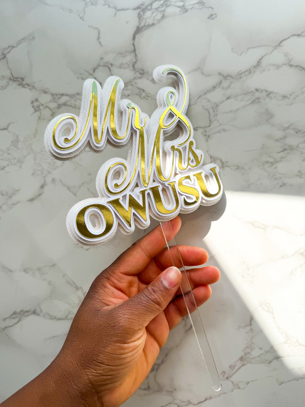 A hand holding a clear acrylic cake topper with gold foil script text reading 'Mr & Mrs OWUSU' against a white and gray marble background.